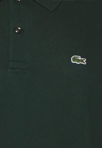 Polo shirt vert foncé en coton texturé, avec un col classique, deux boutons et un petit logo crocodile brodé sur la poitrine.