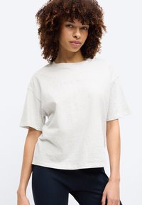 Νεαρή γυναίκα με σγουρά μαλλιά, φορώντας άνετο ανοιχτό γκρι μπλουζάκι και σκούρο μπλε παντελόνι, στέκεται μπροστά σε απλό φωτεινό φόντο.