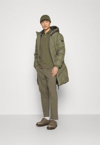 Olivengrøn polstret jakke med hætte, olivenfarvet polo-shirt, khaki bukser og brune sko. Blød tekstur og afslappet pasform.