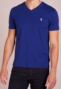 V-ringad t-shirt för män i kunglig blå bomull. Har korta ärmar och en liten gul logotyp på vänster bröst. Slät textur.