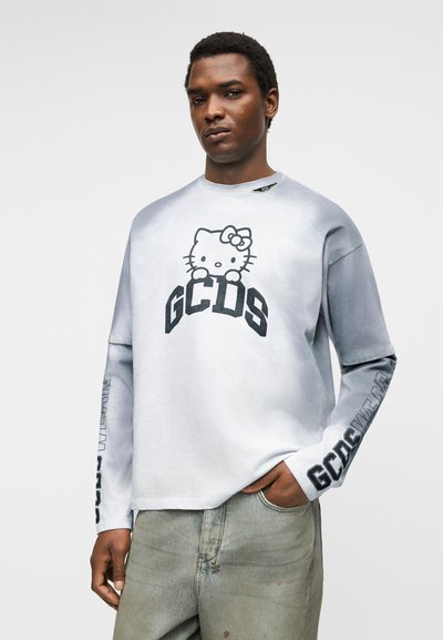 Mies, joka käyttää valko-harmaata pitkähihasta paitaa, jossa on Hello Kitty ja GCDS-logo, yhdistettynä vaaleanharmaisiin farkkuihin, poseeraa käsi taskussa.