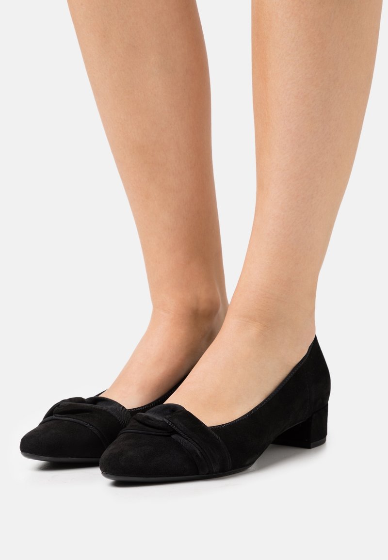 Chaussures basses en daim noir avec un nœud décoratif portées pieds nus sur un fond clair uni.