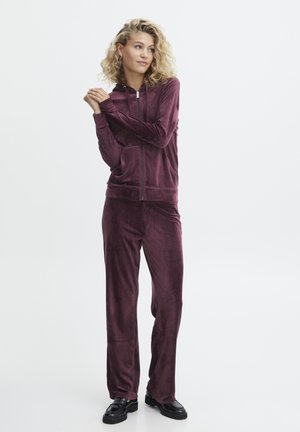 Fluwelen trainingspak in een diepe bordeauxrode kleur, met een ritskap en wijde broek, gecombineerd met zwarte schoenen. Gladde textuur overal.