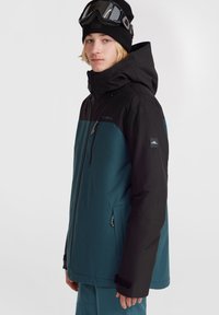 Chaqueta impermeable negra y azul verdoso con capucha, cremallera frontal y bolsillos laterales. Cuenta con puños ajustables con velcro y un logotipo de marca prominente.