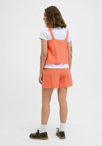 Oranžový kockovaný crop top a zodpovedajúce šortky vyrobené z textúrovanej látky, s širokými ramienkami a uvoľneným strihom. Nosené s bielymi ponožkami a tmavými topánkami.
