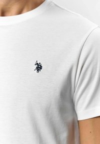 Bijela pamučna majica s okruglim izrezom. Mali mornarski plavi izvezeni logo igrača polo-a smješten je na lijevom dijelu prsnog koša.