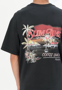 Svart bomull T-skjorte med et fargerikt grafisk motiv av en tropisk scene med palmetrær, vann og teksten "SLIM JIMS COFFEE SHACK PANAMA."