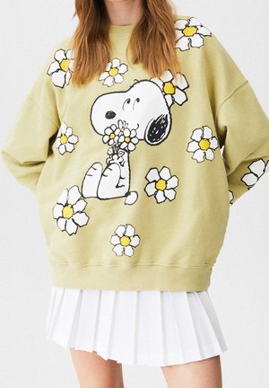 Osoba nosząca jasny zielony sweter z Snoopy'm trzymającym kwiaty i rozrzuconymi białymi i żółtymi stokrotkami, w połączeniu z białą plisowaną spódnicą.