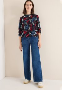 Mønstret bluse i flere farver med lange ærmer, kombineret med high-waisted brede blå jeans og hvide sko med beige såler.