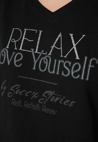 Camiseta negra de cuello en V con adornos de strass, que presenta letras en blanco y gris que dicen: "RELÁJATE, Ámate a ti mismo" y "por Soccer Stories."