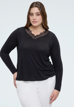 Kvinde iført en sort top med lange ærmer og blonder ved V-halsen samt hvide bukser, stående med den ene hånd på hoften mod en lys baggrund.