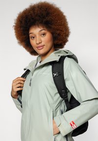 Veste imperméable vert clair avec fermeture éclair à l'avant, capuche ajustable et poignets à velcro. Comprend un sac à dos noir avec des bretelles rembourrées.