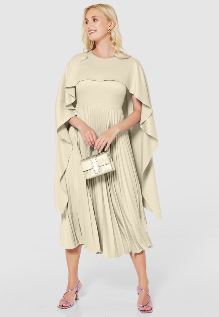Beige geplooide jurk met een cape overlay, gemaakt van lichtgewicht stof. Heeft een getailleerde lijfje en korte mouwen. Vergezeld van een kleine, zilveren handtas.