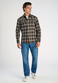 Karierter Flanellhemd in Schwarz und Beige, mit zwei Brusttaschen und Knopfverschluss, kombiniert mit blauen Jeans und weißen Sneakers.