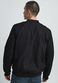 Crna bomber jakna s glatkom, mat završnom obradom. Ima rebraste manžete i pojas, te džep sa zatvaračem na rukavu. Opušteni kroj.