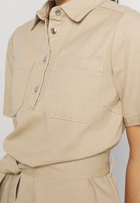 Chemise beige à manches courtes avec col, boutons sur le devant, deux poches poitrine, et ceinture assortie en tissu nouée à la taille.