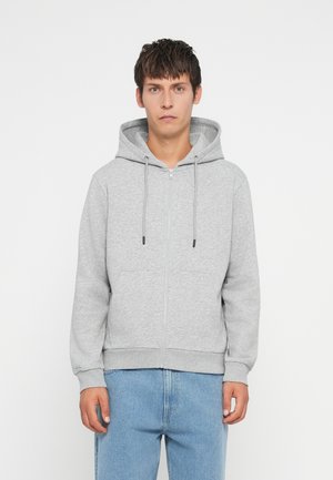Grå glidelås hettegenser laget av mykt stoff, med snor i hetta, frontlommer og ribbestrikkede mansjetter og kant. Bært over lyseblå jeans.