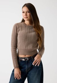 Brauner Strickpullover mit gerippter Textur, rundem Ausschnitt und taillierter Passform, kombiniert mit lockeren Jeans und einem braunen Gürtel mit einer metallischen Schnalle.