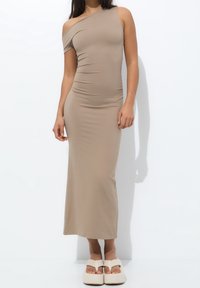 Robe beige longueur genou à une épaule, en tissu extensible avec texture froncée. Portée avec des sandales compensées de couleur claire.