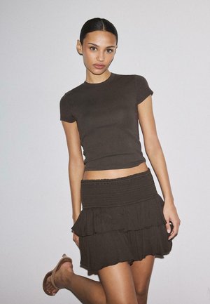 Donna con capelli lisci che indossa una maglietta a maniche corte scura aderente, gonna corta a balze e sandali, posando contro uno sfondo chiaro e semplice.