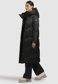 Schwarzer, langer Daunenmantel mit Chevron-Muster, hohem Kragen, seitlichen Reißverschlüssen und Logodetails, kombiniert mit beigen Stiefeln mit dickem Absatz und dunklen Hosen.