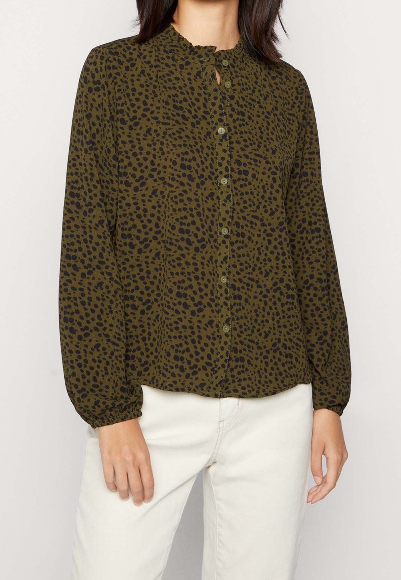 Femme portant un chemisier vert olive à manches longues avec un motif de points noirs irréguliers et un pantalon blanc, debout devant un fond uni.