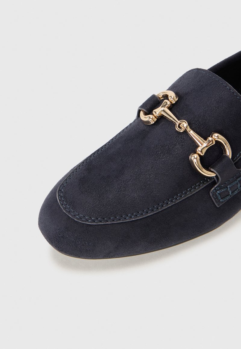 Mocasin de ante en azul marino con punta redondeada, que presenta un detalle en forma de estribo en tono dorado y costuras en contraste a lo largo del borde.