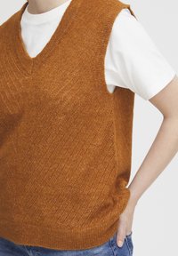 Strickweste in lebhaftem orange-braun, mit V-Ausschnitt, diagonaler Struktur und geripptem Saum. Über einem weißen T-Shirt getragen.