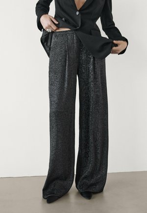 Zwart, metallic van textuur gemaakte wijde broek met plooien, met een hoge taille en een ontspannen pasvorm. G gedragen met een op maat gemaakte zwarte blazer.