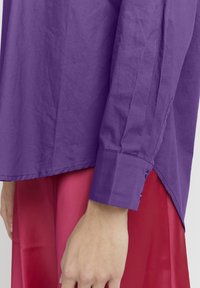 Langärmliges Hemd aus glänzendem, lebhaft lila Baumwollstoff mit abgerundetem Saum und Knopfmanschetten, das eine glatte Textur und einen leicht schimmernden Effekt aufweist.