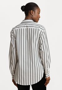 Cămașă cu nasturi din material alb cu dungi verticale negre. Prezintă un guler clasic, mâneci lungi și o margine rotunjită.