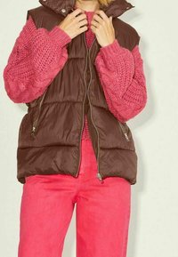 Gilet rembourré marron avec poches zippées, associé à un pull en tricot rose texturé. Le mannequin porte un pantalon rose vif.