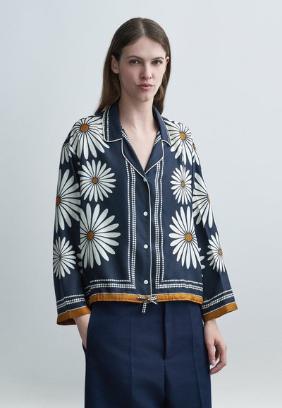 Femme portant un chemisier bleu marine à boutons avec de grands motifs marguerite blancs et une bordure moutarde, associée à un pantalon bleu foncé, debout devant un fond uni.