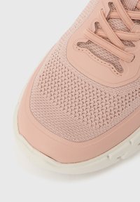 Ανοιχτό ροζ πλεκτό αθλητικό παπούτσι με ροδακινί συνθετικές επικαλύψεις και κορδόνια, με λευκή υφή σόλα σε λευκό φόντο.