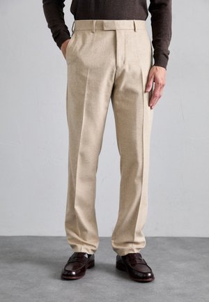 Pantalon en beige tissé avec une coupe ajustée, deux poches avant, des plis simples et une texture lisse, associé à des chaussures en cuir marron.