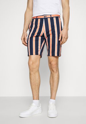 Mann trägt marineblaue Shorts mit vertikalen orange und weißen Streifen, weiße Socken, weiße Turnschuhe und ein weißes T-Shirt.