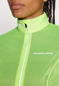 Giacca da ciclismo verde brillante realizzata in materiale leggero. Presenta un colletto alto, accenti riflettenti e il logo 'ENDURA' in bianco.