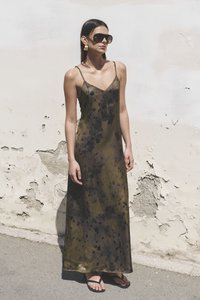 Robe maxi en marron-vert avec des taches noires, en tissu léger. Dispose de bretelles fines et arrive jusqu'aux chevilles, portée avec des tongs.