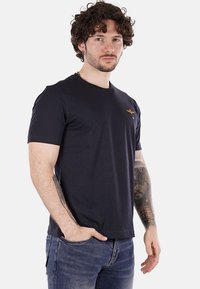 T-shirt blu navy a maniche corte con collo rotondo e un piccolo dettaglio ricamato sul petto. Indossato con jeans di denim blu chiaro.