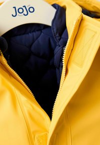 Gul vandtæt jakke med marineblå quilted foer, der har en lynlås og velcro-lukning, hængende på en bøjle.