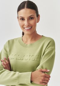 Sudadera verde con un diseño texturizado de "BALANCE" en la parte frontal, cuello redondo y puños acanalados. El material parece suave y cómodo.