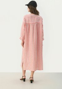 Gestreiftes Maxikleid aus rotem und weißem Leinen, mit langen Ärmeln, lockerer Passform, seitlichen Schlitzen und knöpfbaren Manschetten. Schwarze Kappe und Sandalen mit Blockabsatz.