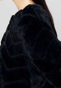 Personne portant un vêtement à manches longues noir, en tissu pelucheux et texturé, avec de longs cheveux foncés visibles sur une épaule, sur un fond uni.