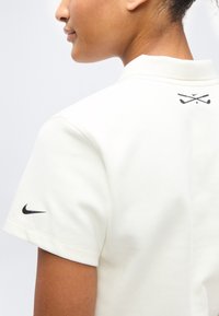 Personne portant un polo blanc avec un swoosh Nike noir sur la manche et des clubs de golf croisés avec une balle de golf sur le haut du dos.