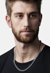Silberne Kettenhalskette mit runden Gliedern, über einem schwarzen T-Shirt getragen. Nahaufnahme der Gesichtszüge mit kurzem Bart und braunen Augen.