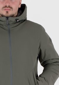 Groene capuchonjas met een gladde textuur, een centrale ritssluiting en zijzakken. Heeft subtiele branding op de mouw.