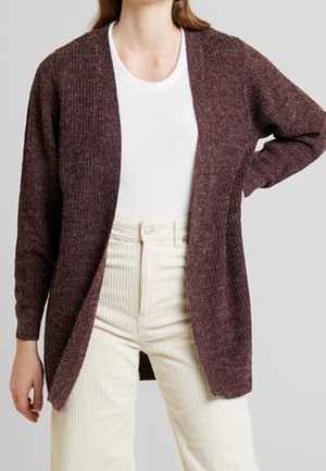 Kobieta w białej koszulce, kremowych sztruksowych spodniach oraz otwartym, prążkowanym ciemnofioletowym kardiganie z przodu.