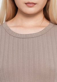 Lèvres et partie inférieure du visage d'une personne à la peau claire et aux cheveux blonds portant un haut en maille côtelée taupe.