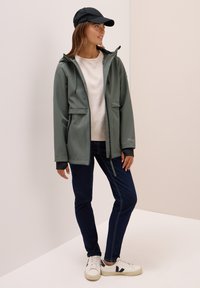 Veste imperméable vert olive avec capuche, deux poches avant, accents noirs sur les poignets, associée à un sweat-shirt blanc et un jean foncé.