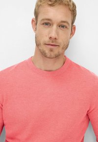 Korallfarbener Strickpullover mit Rundhalsausschnitt und strukturierter Oberfläche. Der Stoff wirkt weich und präsentiert eine klassische Passform sowie dezente Designdetails.